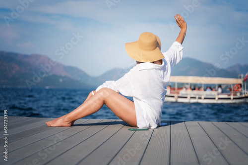 Summer sea resorts concept. A woman in a hat and a white shirt is waving her hand to a pleasure boat on the sea from the shore
