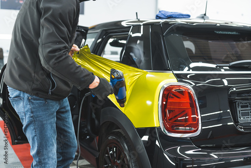 Unrecognisable car detailing specialist using heat gun to glue colourful car wrap to car paint. Indoor medium shot. High quality photo