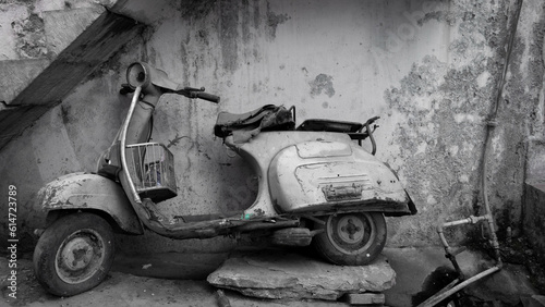 Black and white photography of a scooter abandoned against a stone wall with rocks around