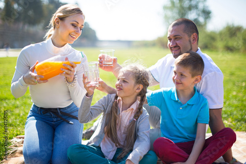 mother pours orange juice into glasses to husband, daughter and son at picnic in park