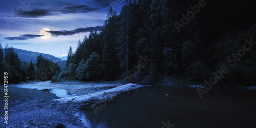 countryside landscape with mountain river at night. trees along the rocky shore and forest on the hill in full moon light