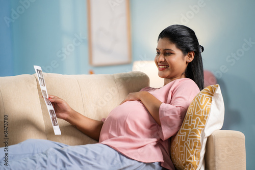 Happy indian pregnant woman on sofa looking ultrasound scan report by holding tummy at home - concept of emotional connection, maternal bonding and expectant motherhood