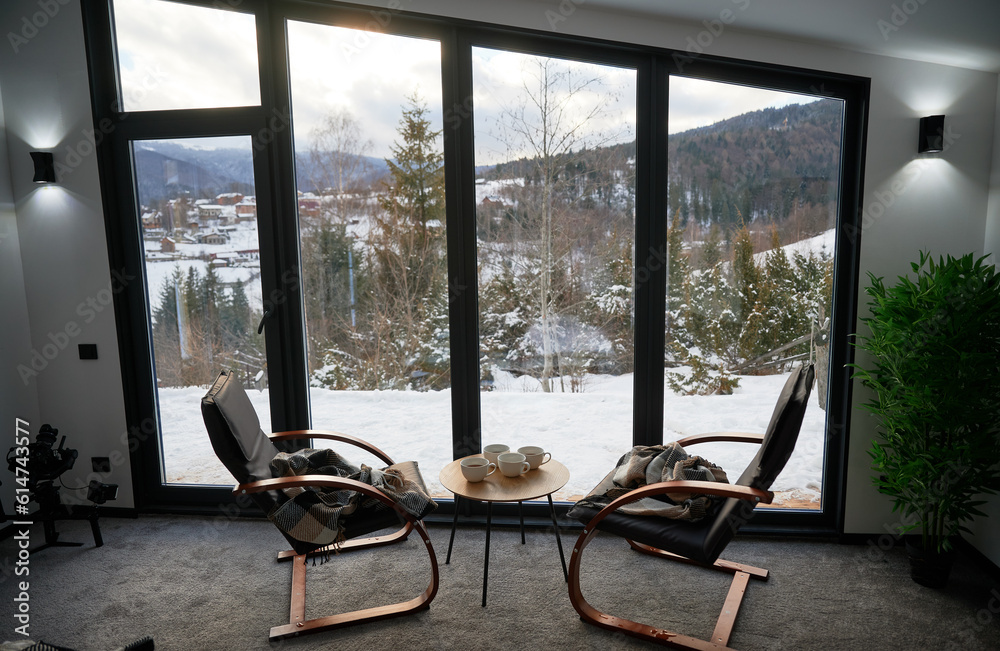 Cozy room with small coffee table and armchairs in front of large panoramic window. Front view of two wooden chairs and table against wide window with snowy mountain landscape. Concept of holidays.