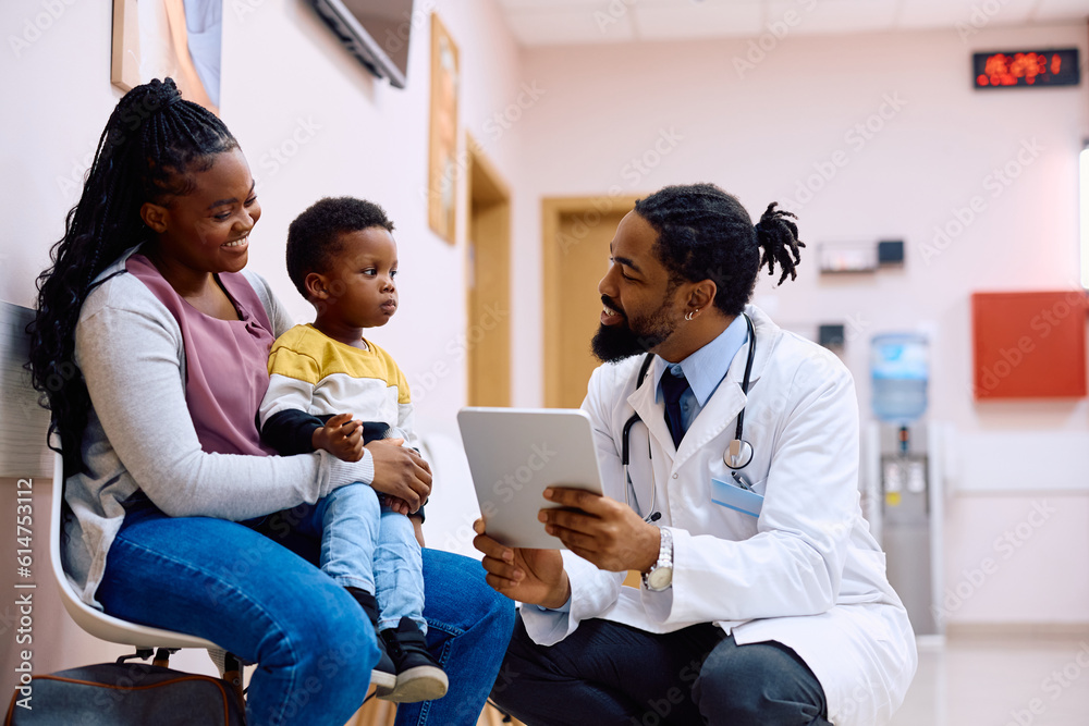 Happy black doctor using touchpad while talking to mother and her son in waiting room at clinic. Happy black doctor using touchpad while talking to mother and her son in waiting room at clinic.