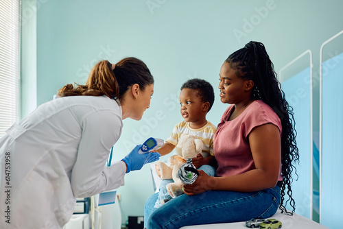Female doctor using infrared thermometer while communicating with black kid and his mother at medical clinic. photo