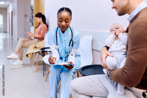 Black pediatric nurse filling medical document of father and his baby at doctor's office.