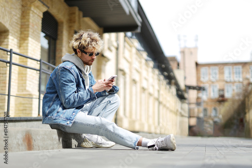 A modern young guy in a denim jacket sits outside and writes a message on the phone.
