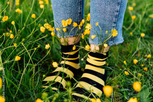 Close up female feet wearing jeans and striped black and yellow socks with flowers inside standing on the green grass of blooming meadow. Concept of bee protection, bloom season, art, creativity. photo