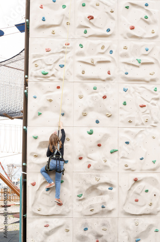 Teenage teen girl grabbing artificial high climbing wall with split ledges. Climbing harness safety sports equipment. Rope amusement city park, obstacle course. Kids children's attraction center