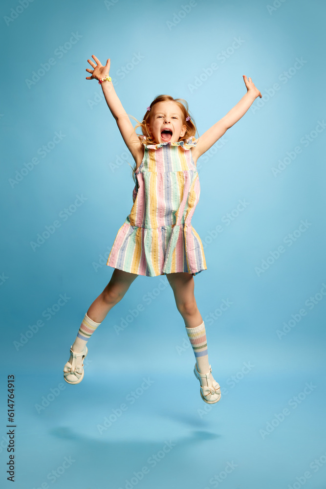 Full-length portrait of beautiful redhead little girl, child in cute striped dress and cheerfully jumping against blue studio background Full-length portrait of beautiful redhead little girl, child in cute striped dress and cheerfully jumping against blue studio background
