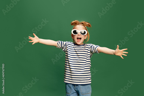 Portrait of emotiona, beautufl little girl, childin striped shirt and sunglasses posing against green studio background photo