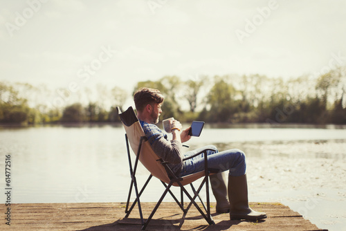 Man drinking coffee and using digital tablet on sunny lakeside dock