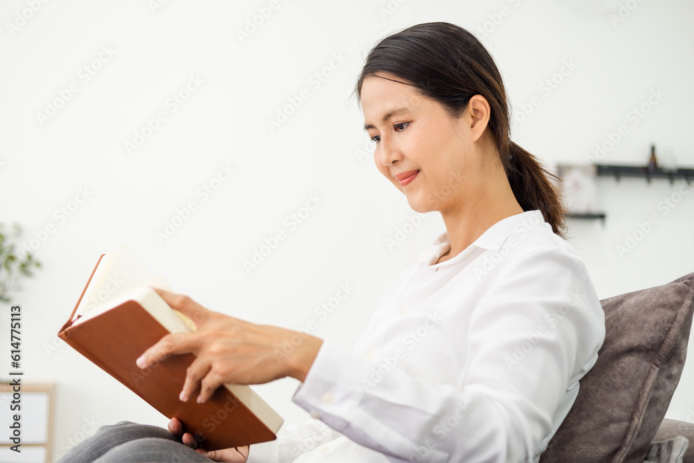 Young beautiful woman reading a book on sofa couch in living room at home . Asian female sitting on textile couch. White wall background, copy space, close up. Young beautiful woman reading a book on sofa couch in living room at home . Asian female sitting on textile couch. White wall background, copy space, close up.
