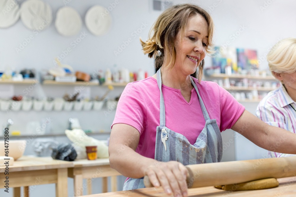 Smiling mature woman rolling clay with rolling pin in pottery studio