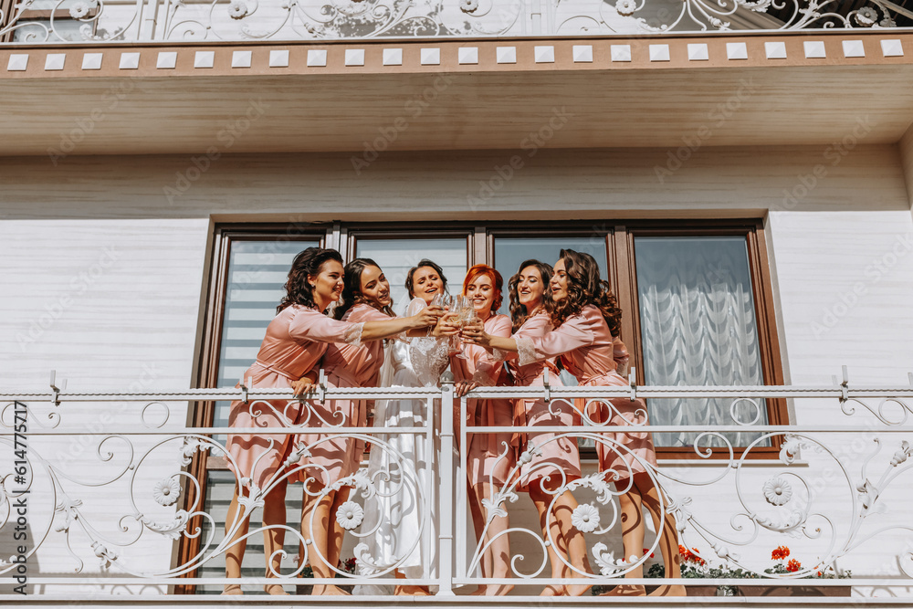 Young bridesmaids are having fun and smiling in pink silk robes drinking champagne at the bride's gazebo. Beautiful women celebrate a bachelorette party, in a gazebo and with champagne.