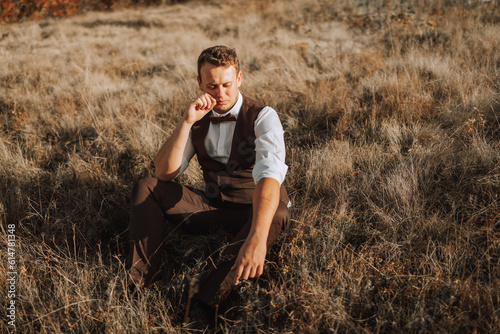 portrait of a stylish groom on a background of dry autumn grass. the concept of a rural wedding in the mountains, happy bohemian newlyweds. man relaxing sitting on the grass