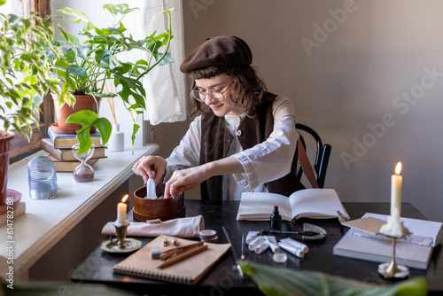 Smiling millennial girl wearing beret choosing paints, sitting at table with painting materials and burning candles, enjoying creating art in cozy home studio. Creative hobbies, artist workplace