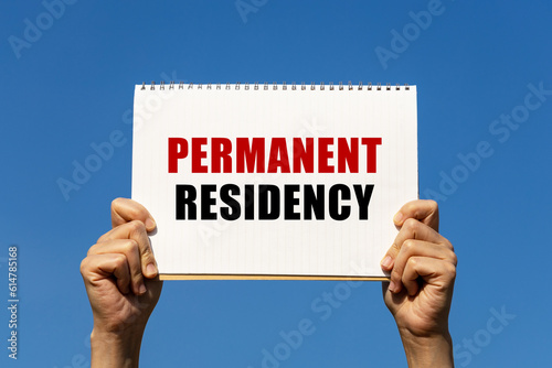 Permanent residency text on notebook paper held by 2 hands with isolated blue sky background. This message can be used as business concept about permanent residency.