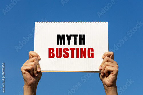 Myth busting text on notebook paper held by 2 hands with isolated blue sky background. This message can be used as business concept about myth busting.