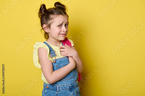 Confused doubtful little preschool child girl, holding her pink backpack, looking at camera with bewildered facial expression, isolated on yellow color background with copy advertising space
