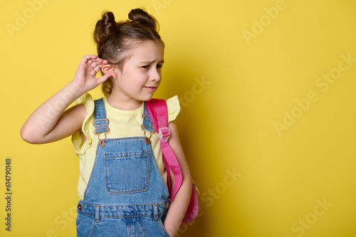 Amazed little girl in denim overalls with a pink backpack, holding her hand to her ear, absorbs the hearing and gossips, expressing bewilderment and surprise, standing isolated on yellow background