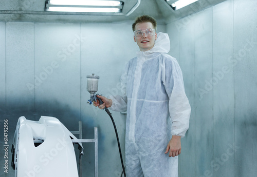 Portrait young man of professional car painter with protective clothing standing next to car in auto paint room and with gun