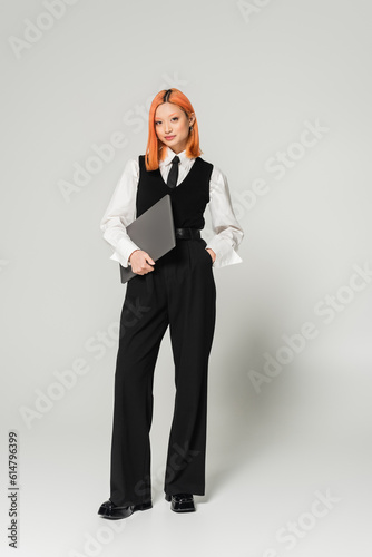 positive and confident freelancer, young asian woman with red colored hair standing with hand in pocket and laptop on grey background, business casual, white shirt, black vest and pants