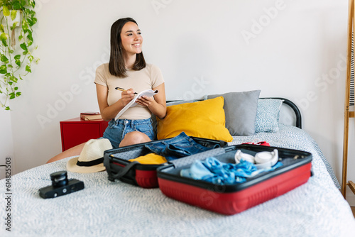 Young beautiful woman sitting on bed packing her suitcase for travel vacation. Smiling female traveler writing planner summer trip on notebook. Summer vacation lifestyle concept.