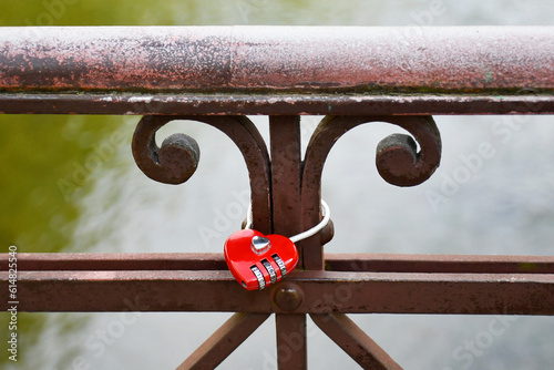 A red lock in the form of a heart hanging on a brown decorative forged element of the bridge railing against on the background of river water