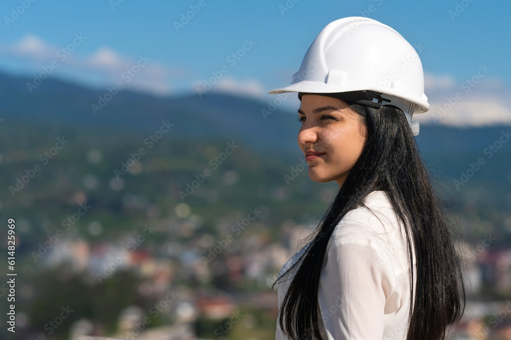 Portrait of a beautiful young female engineer in a white helmet looking forward standing on a construction site on a sunny day against the background of mountains