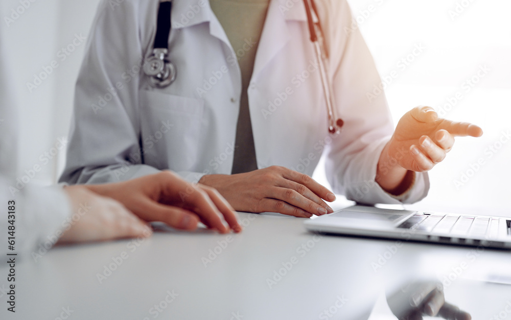 Doctor and patient sitting at the desk in clinic office. The focus is on female physician's hands pointing to laptop computer monitor, close up. Perfect medical service and medicine concept