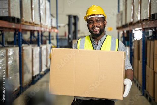 smiling African American male warehouse carrying box parcel and posing looking at the camera.Logistic industry and warehouse concept.Black male workers with Hard Hat working in factory warehouse.