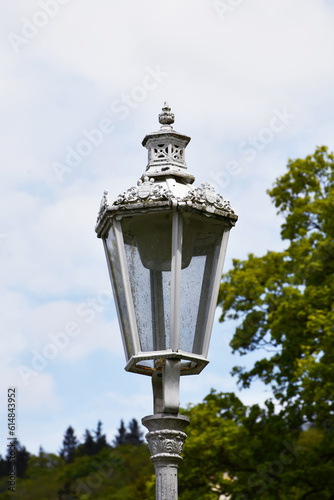 A white lantern with a lot of beautiful elements located in a park on the background of a blue sky and green trees