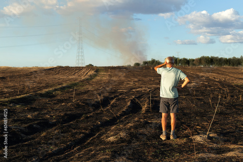 A man with his back turned looks concerned at his burnt field after the fire. He is placed to the right of the image.