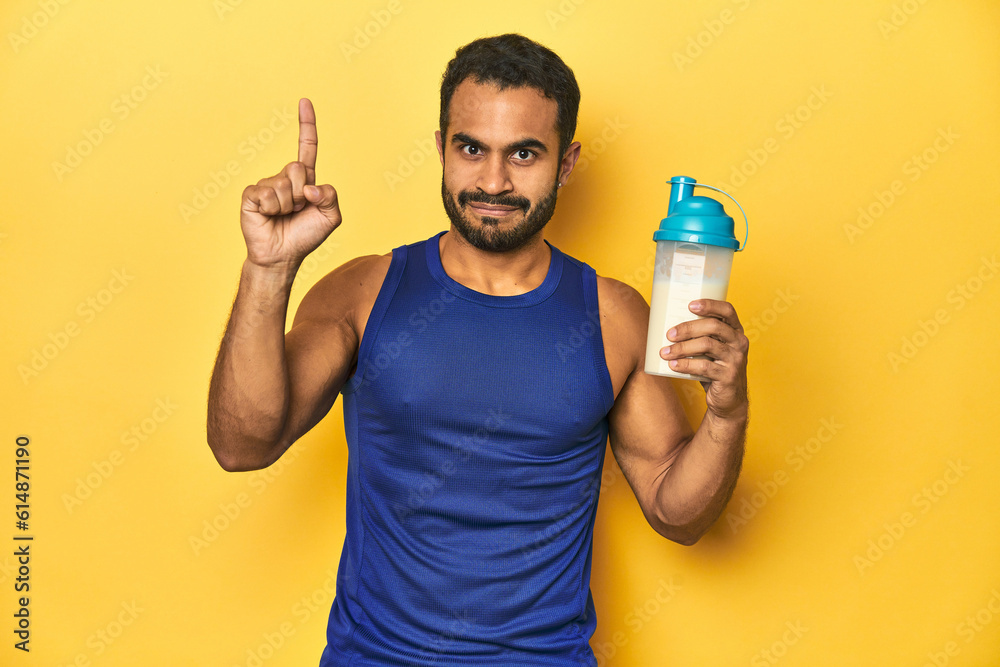 Athletic young Latino man holding a protein shake against a yellow background showing number one with finger. Athletic young Latino man holding a protein shake against a yellow background showing number one with finger.