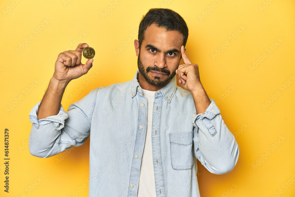 Young Latino man holding a Bitcoin coin, yellow studio background, pointing temple with finger, thinking, focused on a task.