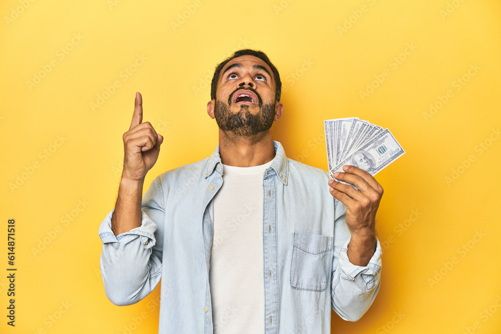 Young Latino man holding a bundle of dollars, yellow studio background, pointing upside with opened mouth. Young Latino man holding a bundle of dollars, yellow studio background, pointing upside with opened mouth.