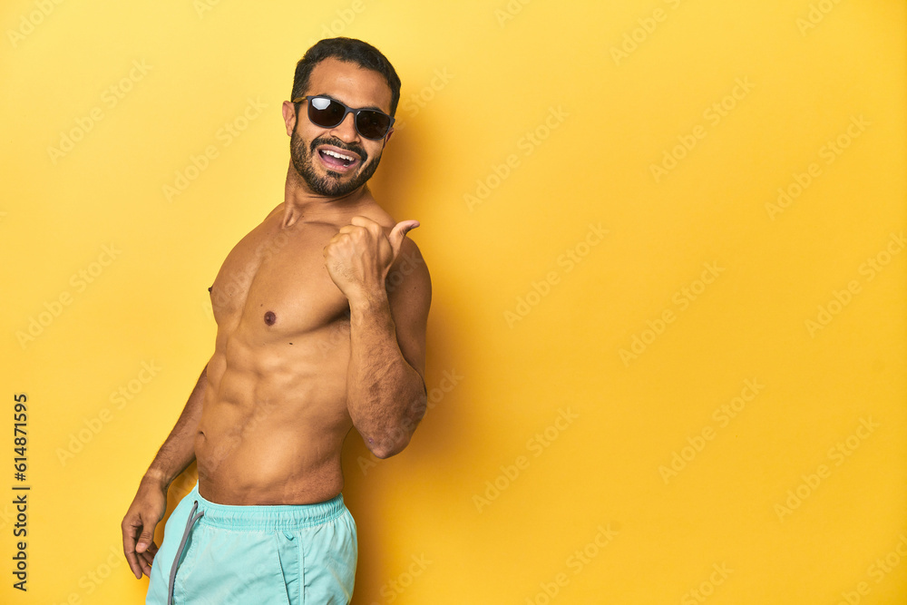 Fit young Latino man in swimwear and sunglasses, yellow studio background, points with thumb finger away, laughing and carefree.