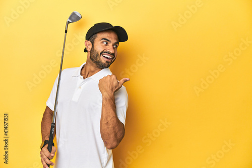 Young Latino golfer with club and cap on a yellow studio background, points with thumb finger away, laughing and carefree.