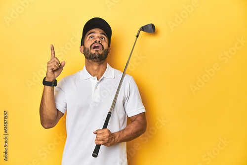 Young Latino golfer with club and cap on a yellow studio background, pointing upside with opened mouth.