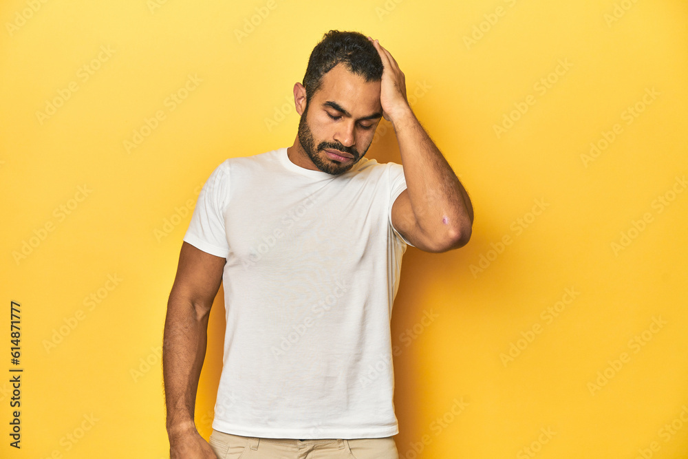 Casual young Latino man against a vibrant yellow studio background, tired and very sleepy keeping hand on head.