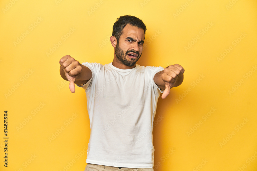 Casual young Latino man against a vibrant yellow studio background, showing thumb down and expressing dislike. Casual young Latino man against a vibrant yellow studio background, showing thumb down and expressing dislike.