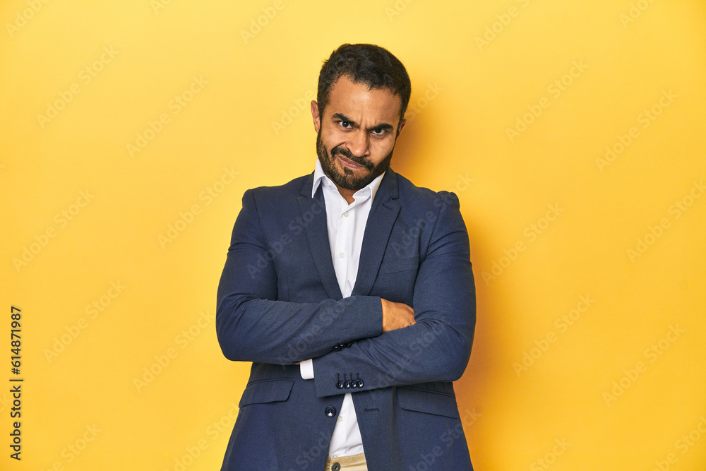 Professional young Latino man in business suit, yellow studio background, frowning face in displeasure, keeps arms folded. Professional young Latino man in business suit, yellow studio background, frowning face in displeasure, keeps arms folded.