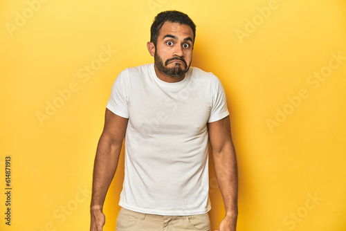 Casual young Latino man against a vibrant yellow studio background, shrugs shoulders and open eyes confused.