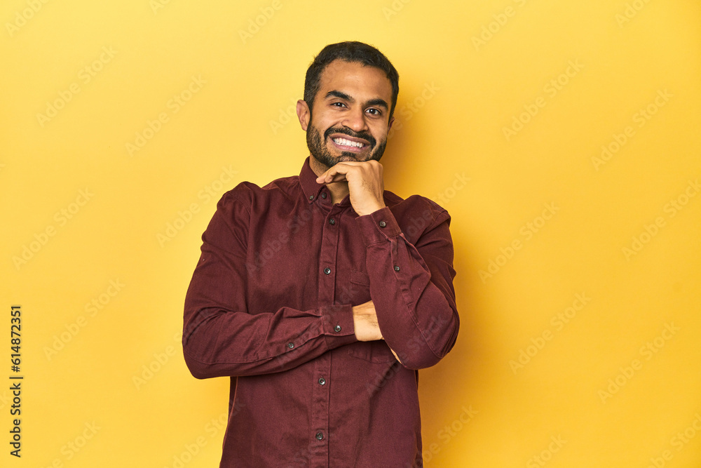 Casual young Latino man against a vibrant yellow studio background, smiling happy and confident, touching chin with hand.
