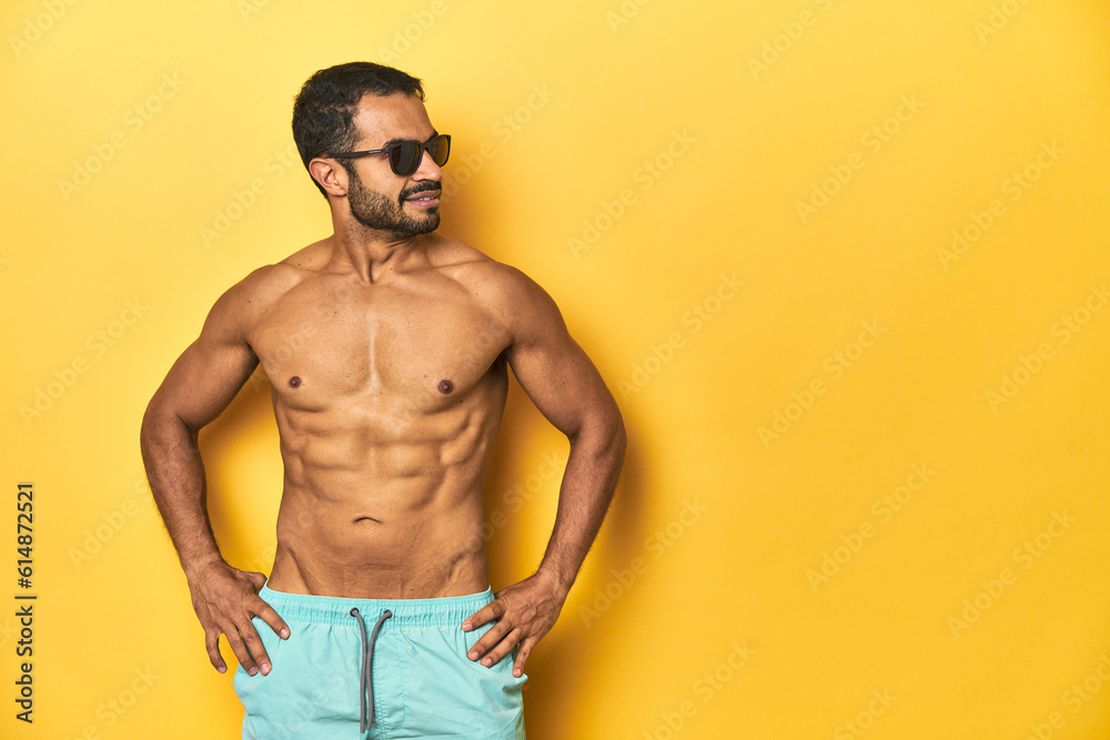 Cheerful muscular Latino man in swimsuit posing with ad space, yellow studio background.