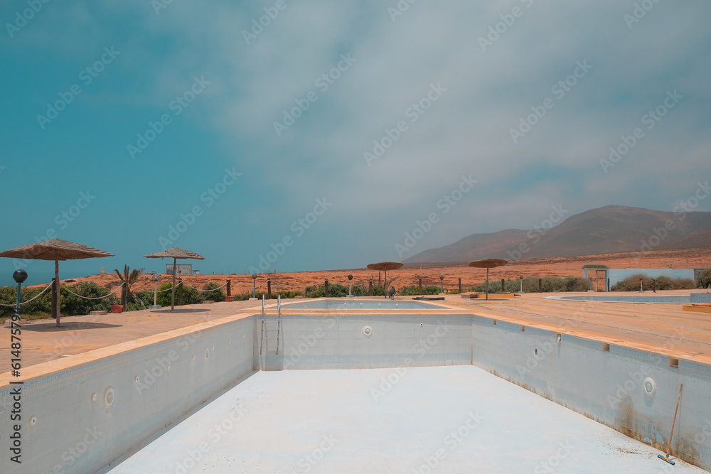 Large empty hotel swimming pool without water in Legzira, Morocco. Blue day sky, huts, mountain views all around. Closed for the season. Bankrupt vacation resort concept. Drained pool background.