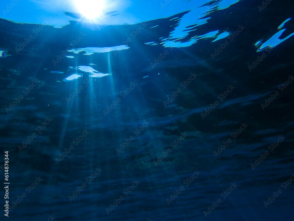 Dark blue ocean surface seen from underwater. Abstract waves underwater and rays of sunlight shining through, Sun light rays undersea deep, Underwater background with sea bottom, Mediterranean sea.