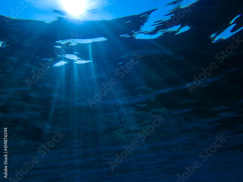Dark blue ocean surface seen from underwater. Abstract waves underwater and rays of sunlight shining through, Sun light rays undersea deep, Underwater background with sea bottom, Mediterranean sea.