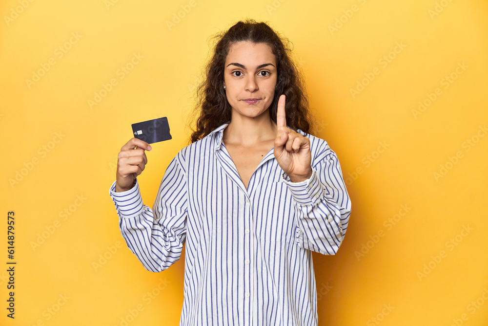 Woman holding a credit card, yellow backdrop, showing number one with finger. Woman holding a credit card, yellow backdrop, showing number one with finger.